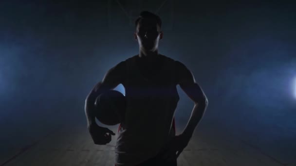 Баскетболист стоит на темной площадке и держит мяч в руках и смотрит в камеру в темноте с подсветкой в замедленной съемке и вокруг дыма — стоковое видео