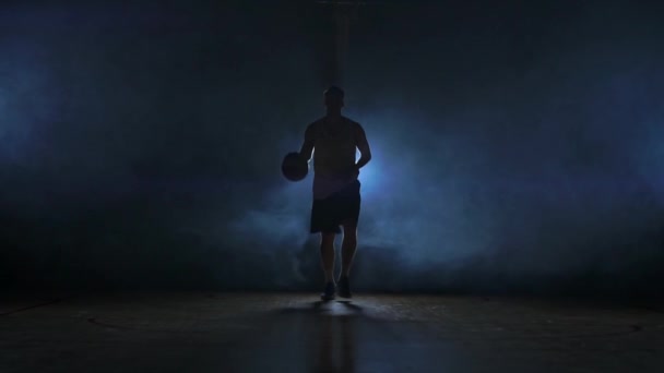 Basketball joueur va directement à la caméra dans une pièce sombre avec un retour rétroéclairé dans la fumée en regardant la caméra au ralenti. Steadicam — Video