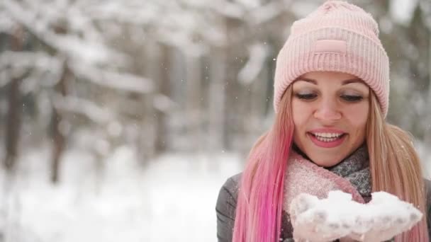 Медленное движение, женщина в куртке шляпа и шарф зимой в лесу держа снег в руках и дуя в камеру бросает снег . — стоковое видео