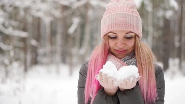 En cámara lenta, una mujer con un sombrero de chaqueta y bufanda en el invierno en el bosque sosteniendo la nieve en sus manos y soplando en la cámara lanza nieve . — Vídeos de Stock