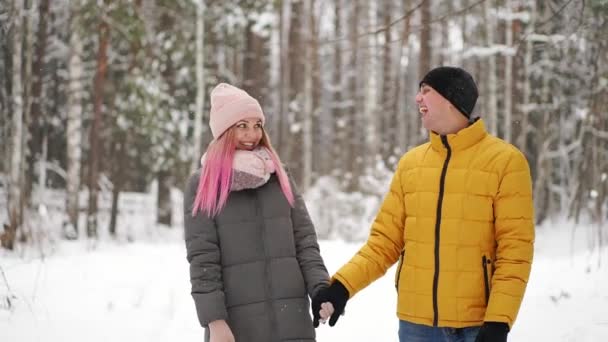 Un hombre con una chaqueta amarilla y una chica con sombrero y bufanda caminan por el bosque de invierno durante una nevada riéndose y sonriéndose en Navidad en cámara lenta — Vídeos de Stock