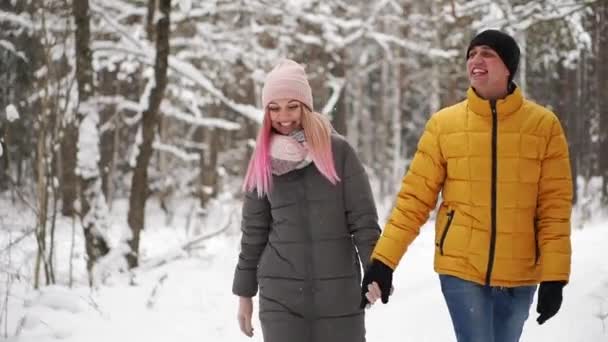 Un uomo in giacca gialla e una ragazza in cappello e sciarpa passeggiano nella foresta invernale durante una nevicata ridendo e sorridendosi a Natale al rallentatore — Video Stock