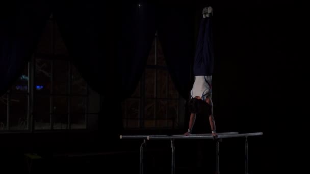 Acrobate gymnaste masculin effectue handstand sur barres parallèles dans une pièce sombre au ralenti partageant un saut périlleux et l'atterrissage sur le sol. Entraînement avant les Jeux Olympiques — Video