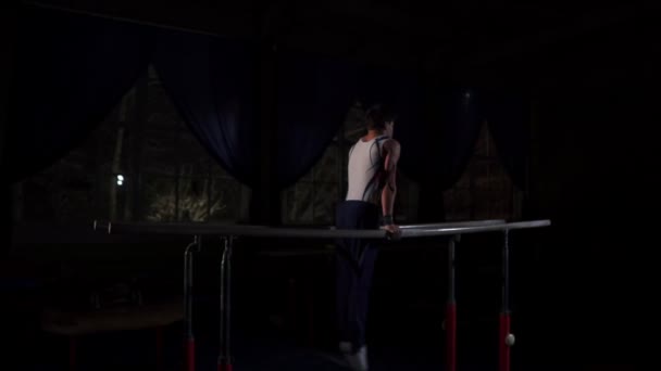 Acrobata de ginasta masculino executa suporte em barras paralelas em uma sala escura em câmera lenta compartilhando um salto mortal e aterrissando no chão. Treinamento antes dos Jogos Olímpicos. Um atleta profissional — Vídeo de Stock