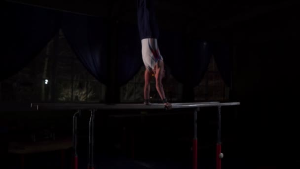 Acrobate gymnaste masculin effectue handstand sur barres parallèles dans une pièce sombre au ralenti — Video