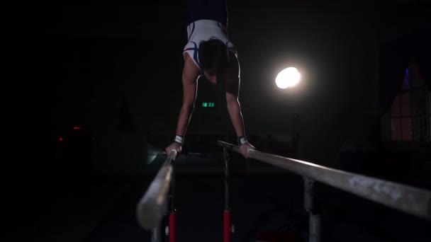 Acrobata de ginasta masculino executa suporte em barras paralelas em uma sala escura em câmera lenta compartilhando um salto mortal e aterrissando no chão. Treinamento antes dos Jogos Olímpicos. Um atleta profissional — Vídeo de Stock