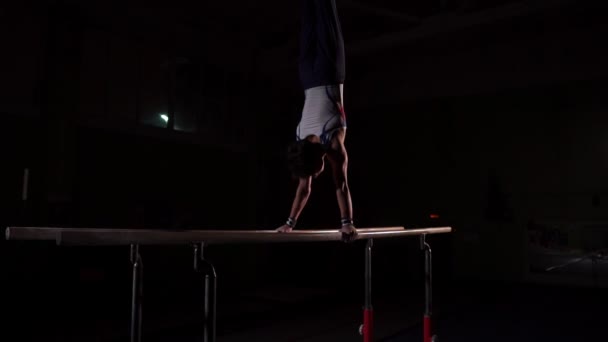 Profesjonalnych gimnastyczka w ciemnym pomieszczeniu w sportowej stoi na raki na poręczach i wykonuje koziołki w zwolnionym tempie. Ciężkich treningów w ramach przygotowań do Igrzysk Olimpijskich — Wideo stockowe