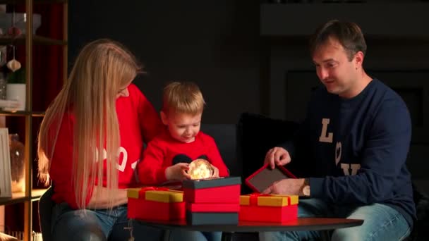 Glückliche Familie, die sich auf die Weihnachtsfeier vorbereitet: Mutter, Vater und Kind lächeln und reden, während sie zu Hause Geschenke öffnen und auf der Couch im Weihnachtsinterieur sitzen. warme und gemütliche Atmosphäre zu Hause — Stockvideo