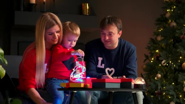 Счастливая семья готовится к празднованию Рождества: мама, папа и ребенок улыбаются и разговаривают, открывая подарки дома, сидя на диване в рождественском интерьере. Теплая и уютная атмосфера дома — стоковое видео