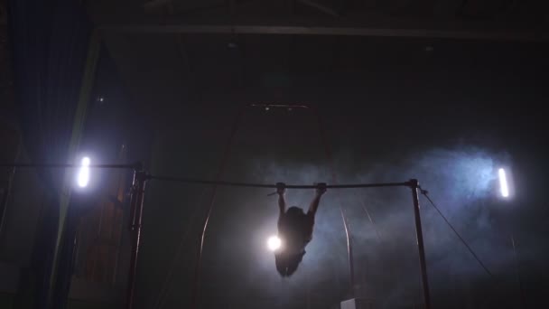 Gymnast on a dark background on the crossbar performs spins and jumps over himself in the air in slow motion and around the smoke — Stock Video