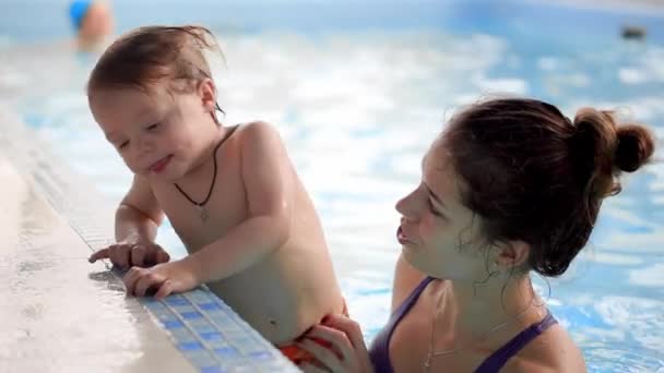 Mãe ajuda o bebê a ficar do lado da piscina durante o treinamento de natação para bebês — Vídeo de Stock