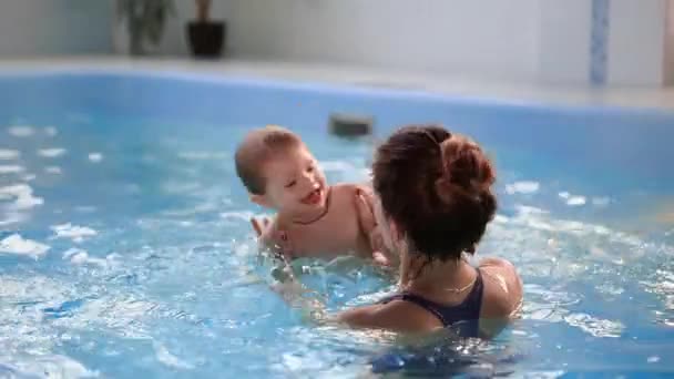 Lindo bebé y su madre teniendo clases de natación en la piscina. La Madre sostiene a su hijo en sus manos y lo abraza. El niñito sonríe alegremente. — Vídeos de Stock