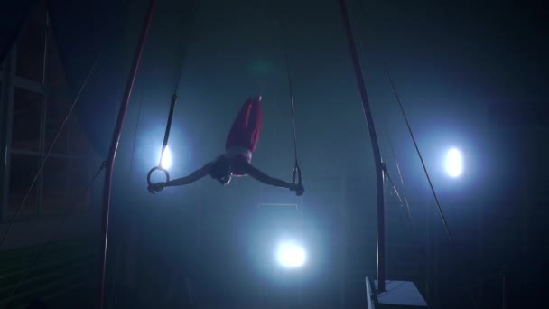 Tornász, sötét háttér előtt áll, kezében gyűrűk segítségével a levegőben. Az olimpiai programban hajtja végre a forgatás — Stock videók