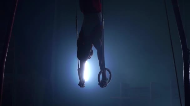 Ginasta em um fundo escuro fica em suas mãos usando anéis no ar. Executa rotação no programa olímpico em câmera lenta 120 fps. anéis de ginástica, ginasta profissional — Vídeo de Stock