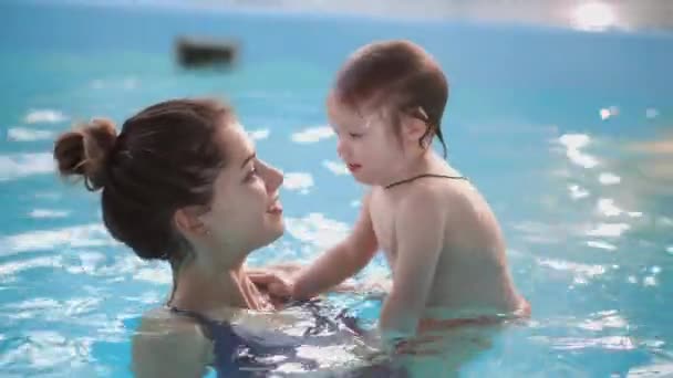 Lindo bebé y su madre teniendo clases de natación en la piscina. La Madre sostiene a su hijo en sus manos y lo abraza. El niñito sonríe alegremente. — Vídeo de stock
