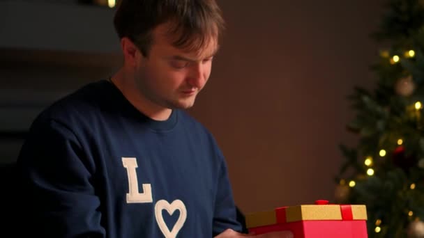 Молодой человек с рождественским подарком в руках открывает его, сидя на диване — стоковое видео