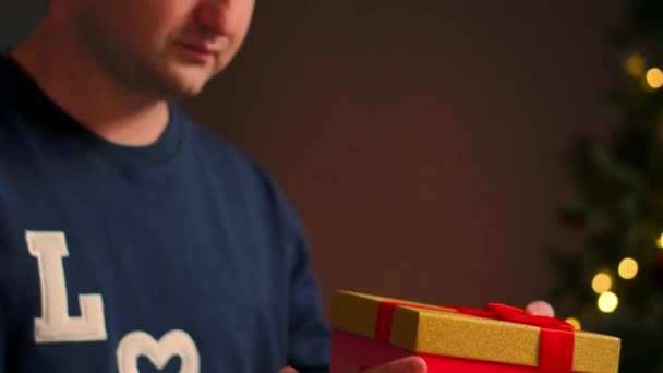 Молодой человек с рождественским подарком в руках открывает его, сидя на диване — стоковое видео