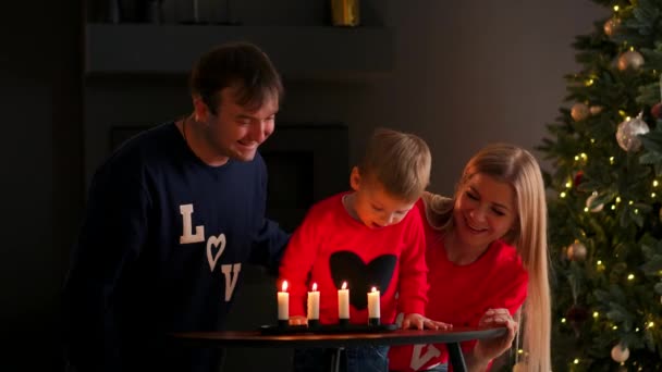 Семья на Рождество сидит под елкой и смотрит, как ребенок задувает свечи и смеется. Мама и папа смеются и улыбаются — стоковое видео