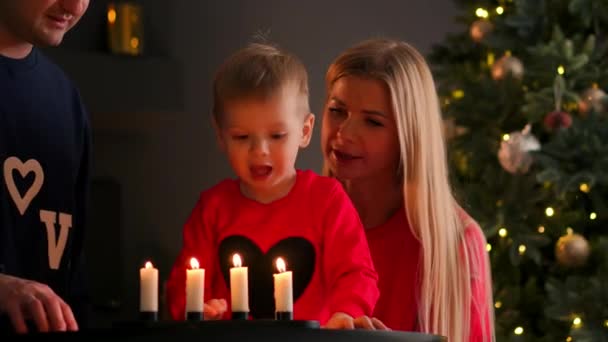 Año nuevo, Navidad, niño y madre y padre junto a la chimenea junto al árbol de Navidad mirando las velas encendidas en los candelabros, la habitación es oscura, la luz de la chimenea y — Vídeos de Stock