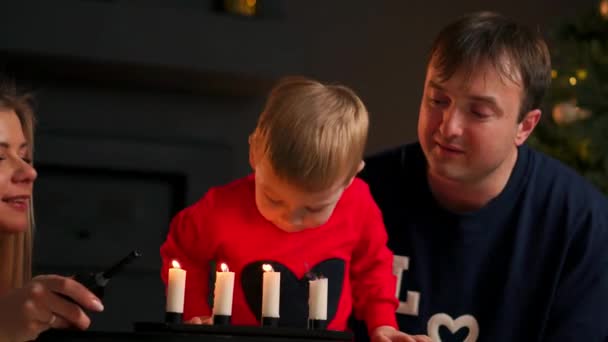 新年, 圣诞节, 男孩和爸爸妈妈在圣诞树旁的壁炉旁看着烛台上燃烧的蜡烛, 房间很黑, 壁炉里的灯光和 — 图库视频影像