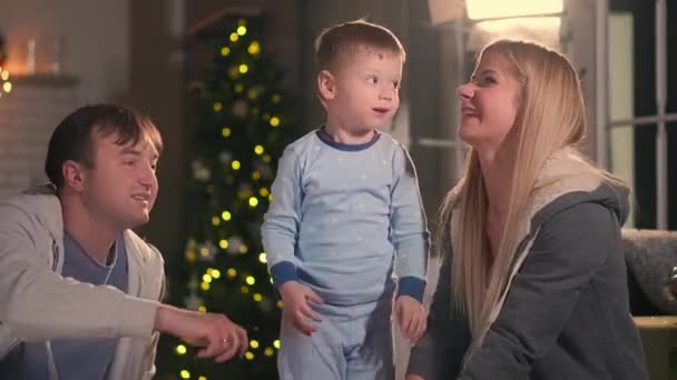 Glückliche Eltern mit ihrem Sohn, der an Weihnachten im Schlafanzug auf dem Bett sitzt und vor Neujahr eine Kissenschlacht spielt — Stockvideo