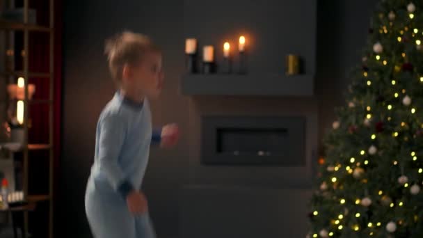 Kleine vrolijke jongen springen op het bed en gooien een kussen op de achtergrond de kerstboom. — Stockvideo