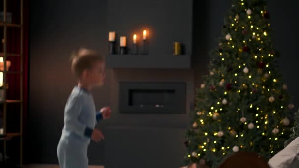 Kleine vrolijke jongen springen op het bed en gooien een kussen op de achtergrond de kerstboom. — Stockvideo