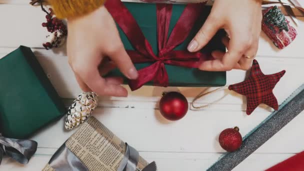 Креативне хобі. Обгортання подарунків. Упаковка сучасних різдвяних подарункових коробок у стильному сірому папері з атласною червоною стрічкою. Вид зверху рук на стіл з білого дерева з гілками ялинки, прикраса — стокове відео
