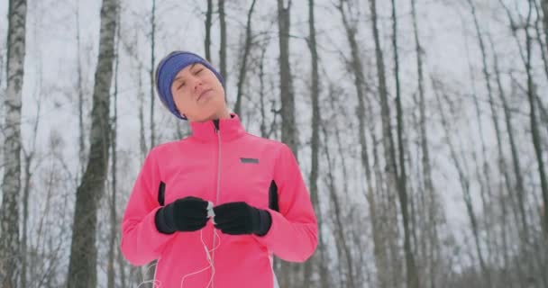 Дівчина перед ранковим зимовим бігом вставляє навушники в вуха і готується пробігти через природний парк у повільному русі. Прослуховування музики під час бігу. Вивчати іноземні мови — стокове відео