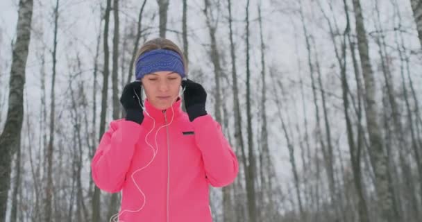 Женщина перед утренней зимней пробежкой вставляет наушники в уши и готовится бегать по природному парку в замедленной съемке. Слушать музыку во время бега. Изучение иностранных языков — стоковое видео