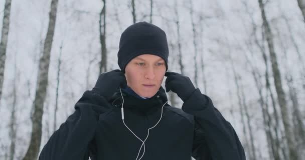 Um jovem numa corrida matinal segura os auscultadores nas mãos e insere-os nos ouvidos antes de correr no parque. Corrida de inverno. Use gadgets modernos e aplicativos de treinamento. Ouça música enquanto — Vídeo de Stock