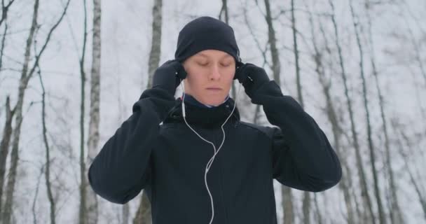 Un jeune homme en jogging matinal tient un casque dans ses mains et l'insère dans ses oreilles avant de courir dans le parc. Course d'hiver. Utilisez des gadgets modernes et des applications de formation. Écouter de la musique pendant — Video