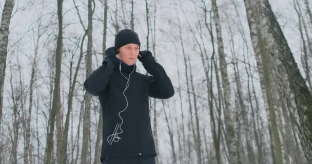 Un joven en una carrera matutina sostiene los auriculares en sus manos e inserta en sus oídos antes de correr en el parque. Carrera de invierno. Utilice aparatos modernos y aplicaciones de entrenamiento. Escuchar música mientras — Vídeos de Stock