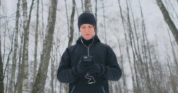 Un jeune homme dans un déclencheur noir en hiver se prépare à faire du jogging dans le parc comprend courir de la musique sur un smartphone et court avec un assistant vocal à travers la forêt faire des affaires et diriger un — Video