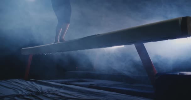 The girl is a professional athlete performs gymnastic acrobatic trick on a beam in backlight and slow motion in sports gymnastic clothing. Smoke and blue. Jump and spin on the balance beam — Stock Video