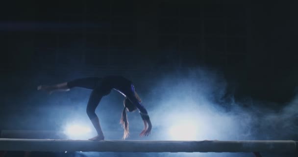 A menina executa um truque em um log na luz de fundo e câmera lenta em roupas de ginástica esportiva. Fumo e azul. Saltar e girar no feixe de equilíbrio — Vídeo de Stock