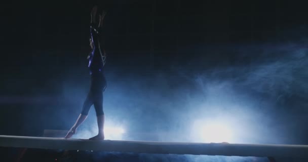 La chica es una atleta profesional realiza truco acrobático gimnástico en un haz de luz de fondo y cámara lenta en ropa deportiva gimnástica. Humo y azul. Saltar y girar en la viga de equilibrio — Vídeos de Stock