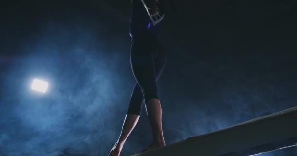 Dziewczyna jest zawodowym sportowcem wykonuje gimnastyki akrobatycznej trick na belkę w podświetlenie i zwolnionym tempie w sportowe gimnastyczne odzież. Dym i niebieski. Skok i spin na belki wagi — Wideo stockowe