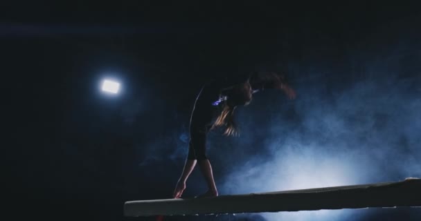 Het meisje is dat een professioneel atleet presteert gymnastische acrobatische truc op een balk in tegenlicht en slow in sport gymnastiek kleding. Rook en blauw. Springen en draaien op de evenwichtsbalk — Stockvideo