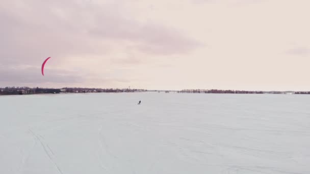 Carrera de competición de kite de nieve de invierno extremo aéreo 4K con diferentes cometas de nieve de colores, esquí, snowboarders sobre el lago de hielo en frente de la ciudad en la ventisca y el clima de nieve del dron — Vídeo de stock