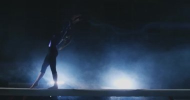 Profesyonel bir atlet bir ışının içinde arka ışık ve jimnastik spor giyim ağır çekimde jimnastik akrobatik numara gerçekleştirir kızdır. Duman ve mavi. Atlama ve denge aleti spin