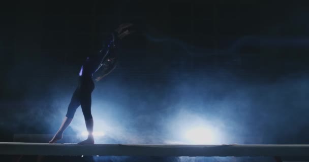 Девушка профессиональная спортсменка выполняет гимнастический акробатический трюк на балке в подсветке и замедленной съемке в спортивной гимнастической одежде. Дым и голубой. Прыжок и вращение на бревне баланса — стоковое видео