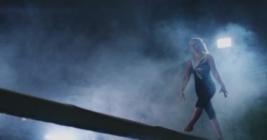 Profesyonel bir atlet bir ışının içinde arka ışık ve jimnastik spor giyim ağır çekimde jimnastik akrobatik numara gerçekleştirir kızdır. Duman ve mavi. Atlama ve denge aleti spin
