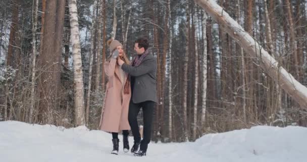 雪に覆われた冬の森、コートやマフラーを着た若い男女が歩くと楽しい。愛するカップルが一緒に過ごすバレンタインの日. — ストック動画
