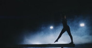 Profesyonel bir atlet bir ışının içinde arka ışık ve jimnastik spor giyim ağır çekimde jimnastik akrobatik numara gerçekleştirir kızdır. Duman ve mavi. Atlama ve denge aleti spin