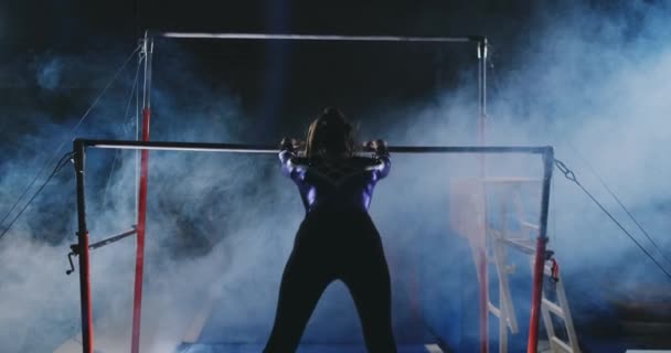 Competitions in gymnastics. A woman walks to the crossbar and performs dips flip-flops in slow motion on a dark background in backlight. Slow motion. — Stock Video