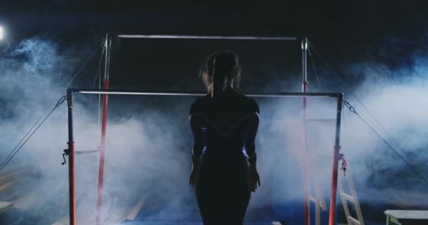 Zawody w gimnastyce. Kobieta idzie do poprzeczkę i wykonuje klapki spadki w zwolnionym tempie na ciemnym tle w podświetlenie. Zwolnionym tempie. — Wideo stockowe