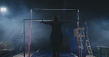 Jimnastik yarışmalarında. Bir kadın için kol demiri yürür ve arka ışık karanlık bir arka plan üzerinde ağır çekimde dips flip-flop gerçekleştirir. Ağır çekim.