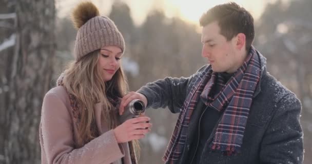 Un couple amoureux homme et femme dans la forêt d'hiver buvant du thé dans un thermos. Homme et femme élégants dans un manteau dans le parc en hiver pour une promenade. Mouvement lent — Video