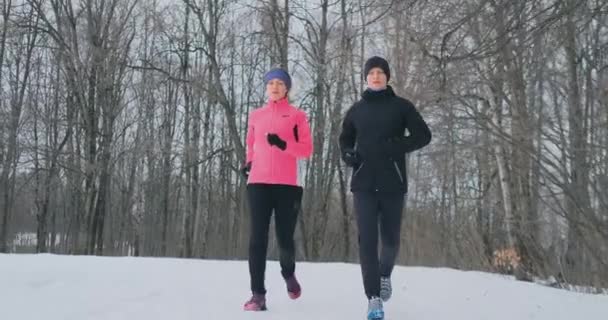 Genç erkek ve kadın kış ormanda koşmak bir sabahı. Bir kadında gevşek bir ceket siyah ceketli adamı bir kış parkı ile çalışıyor. Sağlıklı yaşam mutlu aile. — Stok video